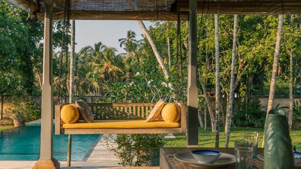 A wooden swing chair with yellow cushions, a table with a green vase, and a pool in the background. - Beautiful Staffed 5 Bedroom Villa, Very Personal Touch Overlooking Paddy Fields (Pitiduwa in Talpe Pattuwa)