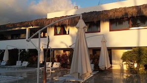 Un espace piscine avec des chaises longues et des parasols, un bâtiment doté d’un toit de chaume et un drapeau.