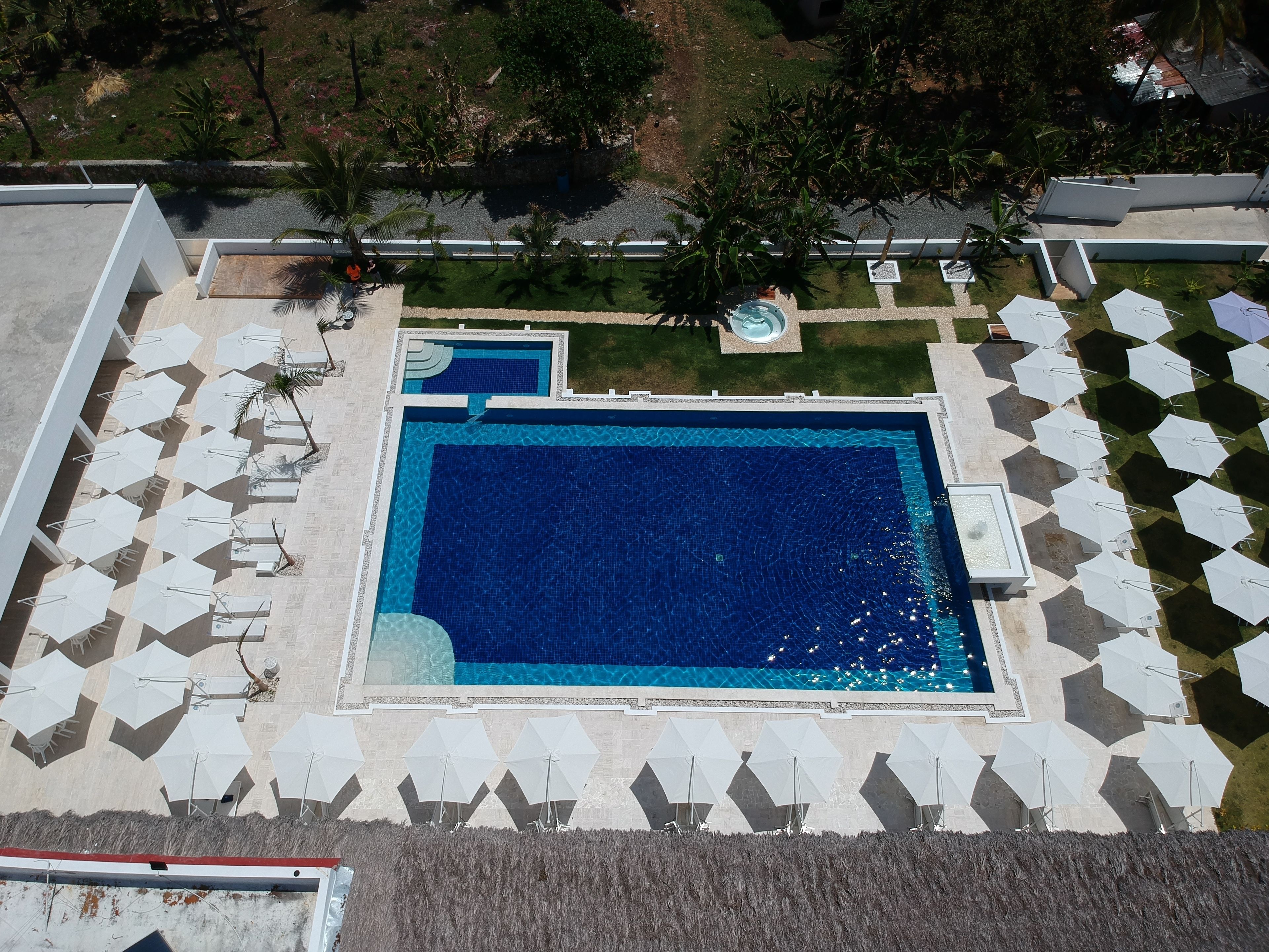 Ein großer rechteckiger Pool mit einem kleineren angrenzenden Pool, umgeben von weißen Sonnenschirmen und Liegestühlen, mit einer angelegten Grünfläche und Bäumen im Hintergrund.