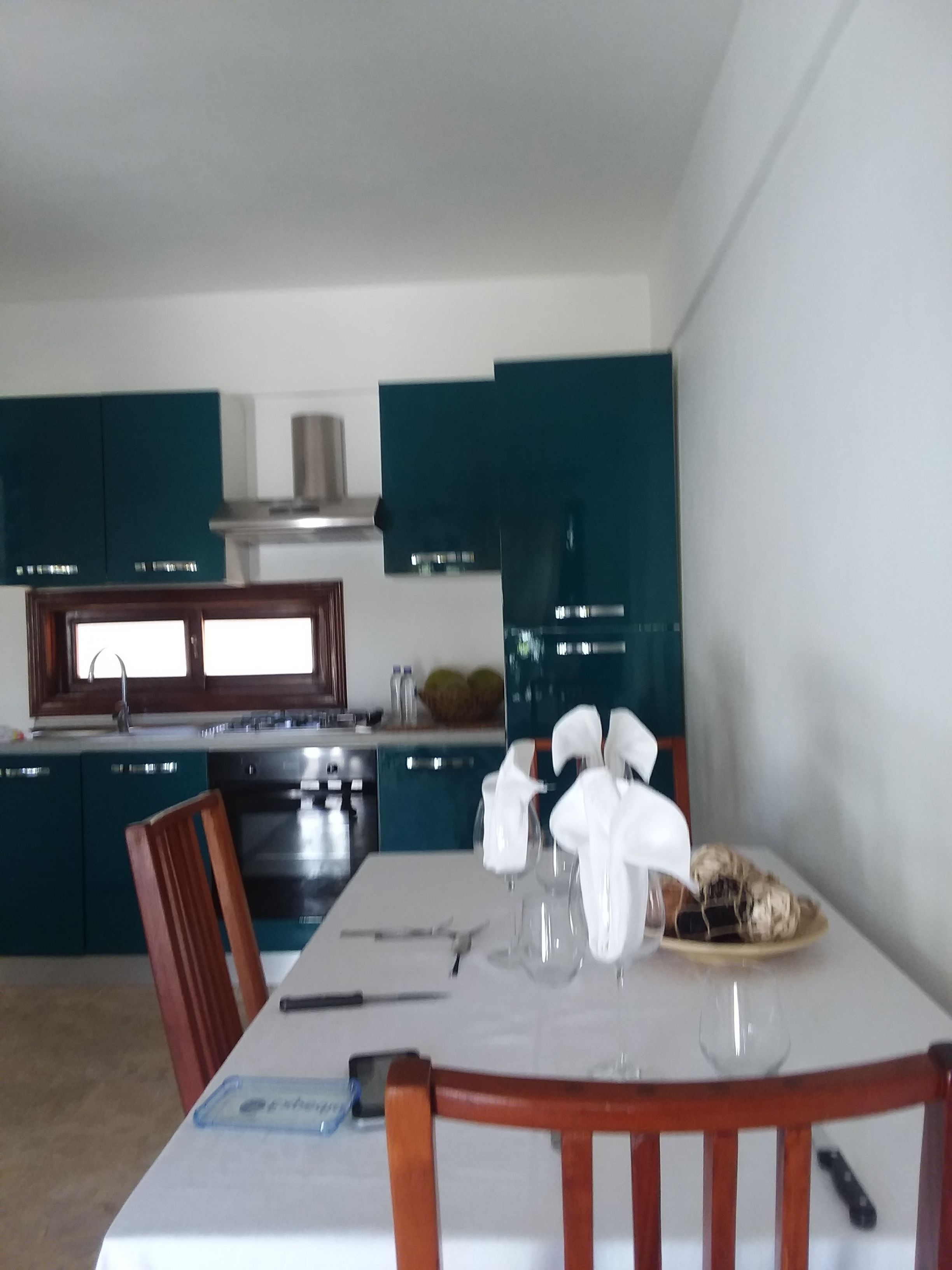 A modern kitchen with teal cabinets, a stainless steel range hood, and a dining area with a white table set for four.