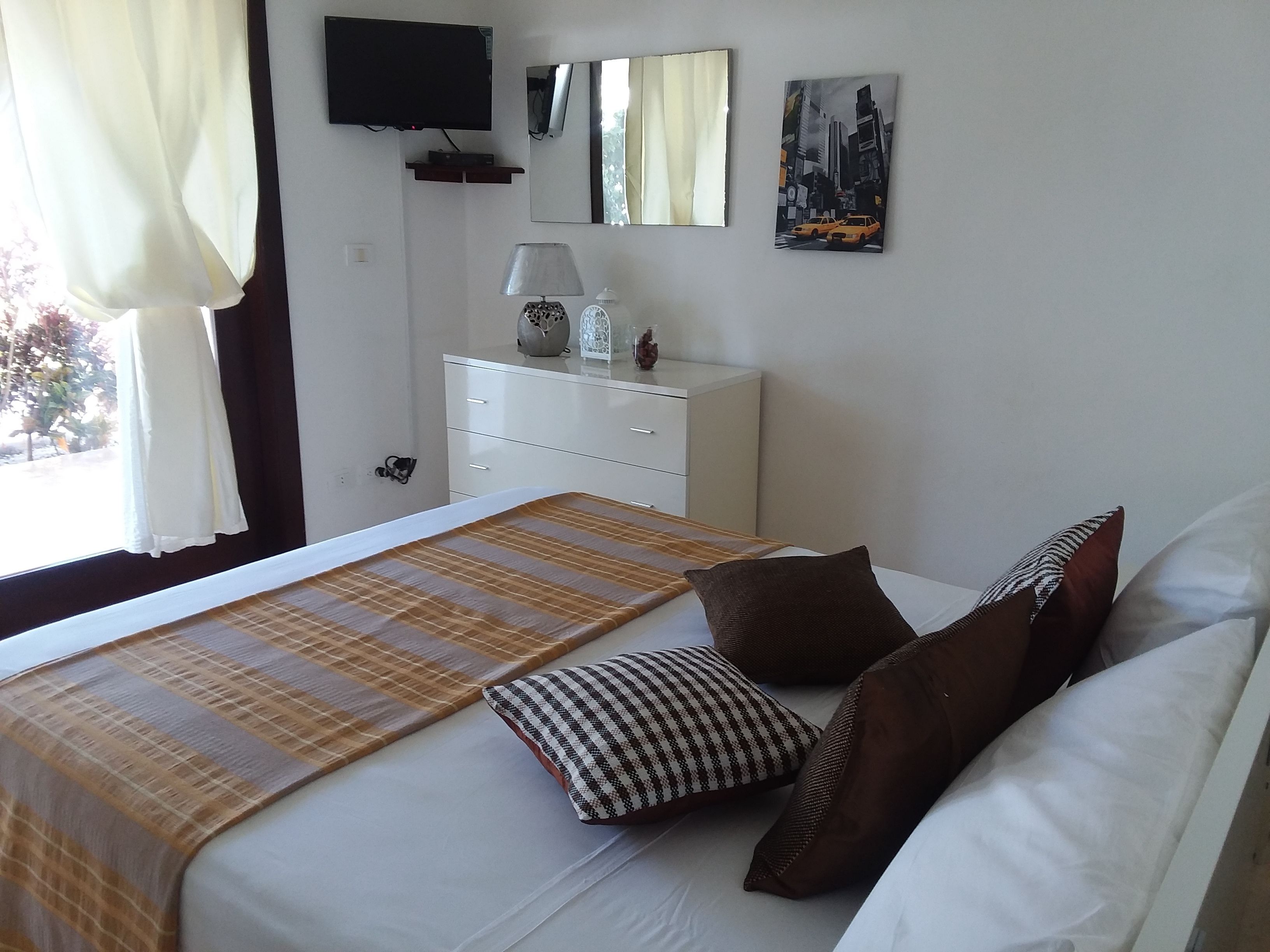 A neatly arranged bedroom with a bed, pillows, a bedside table with a lamp, a dresser, a TV mounted on the wall, and a cityscape painting.