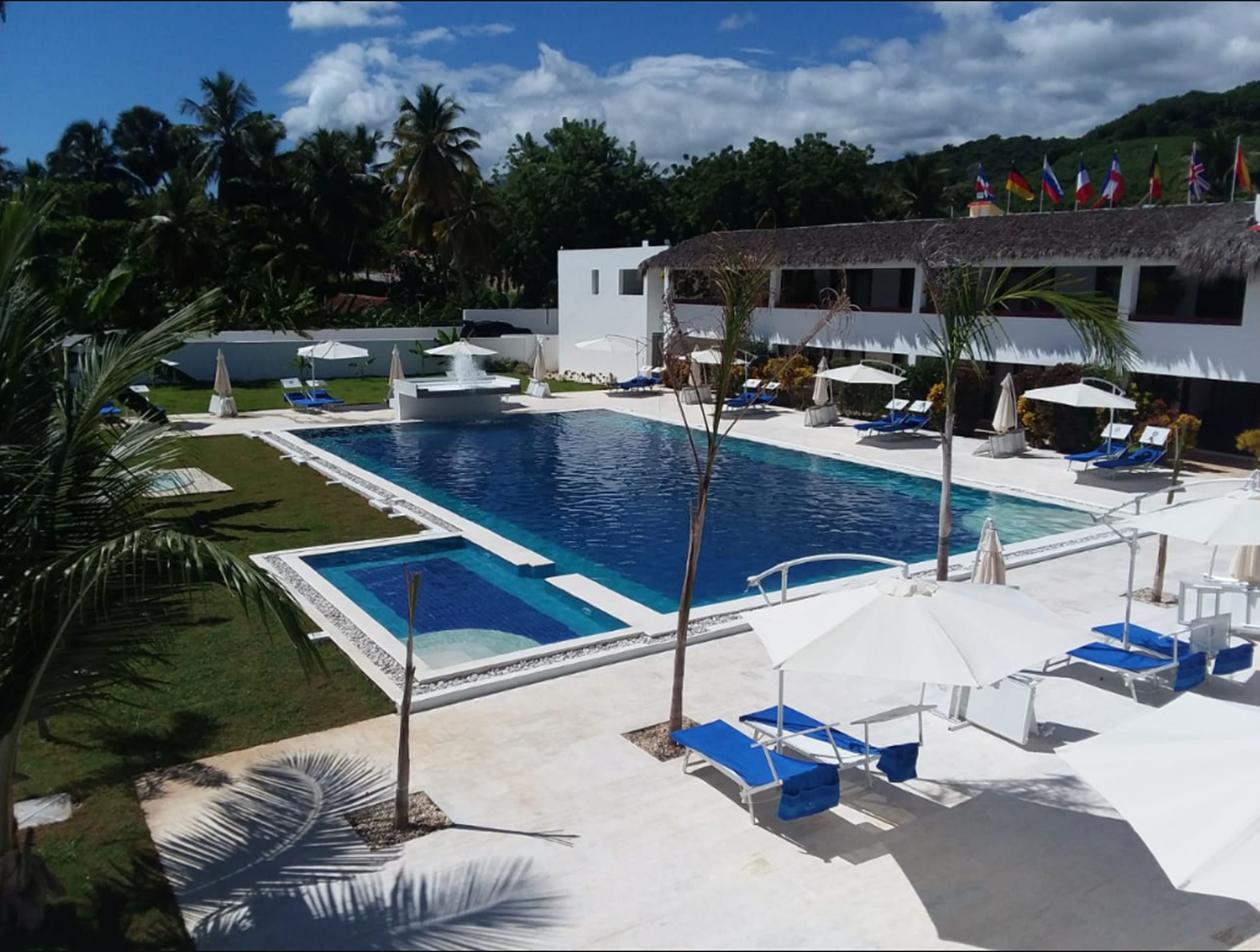 A swimming pool with lounge chairs and umbrellas, surrounded by palm trees and a building with flags.