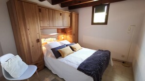 A bedroom with a wooden wardrobe, a bed with a white and blue bedspread, a chair, and a small window.
