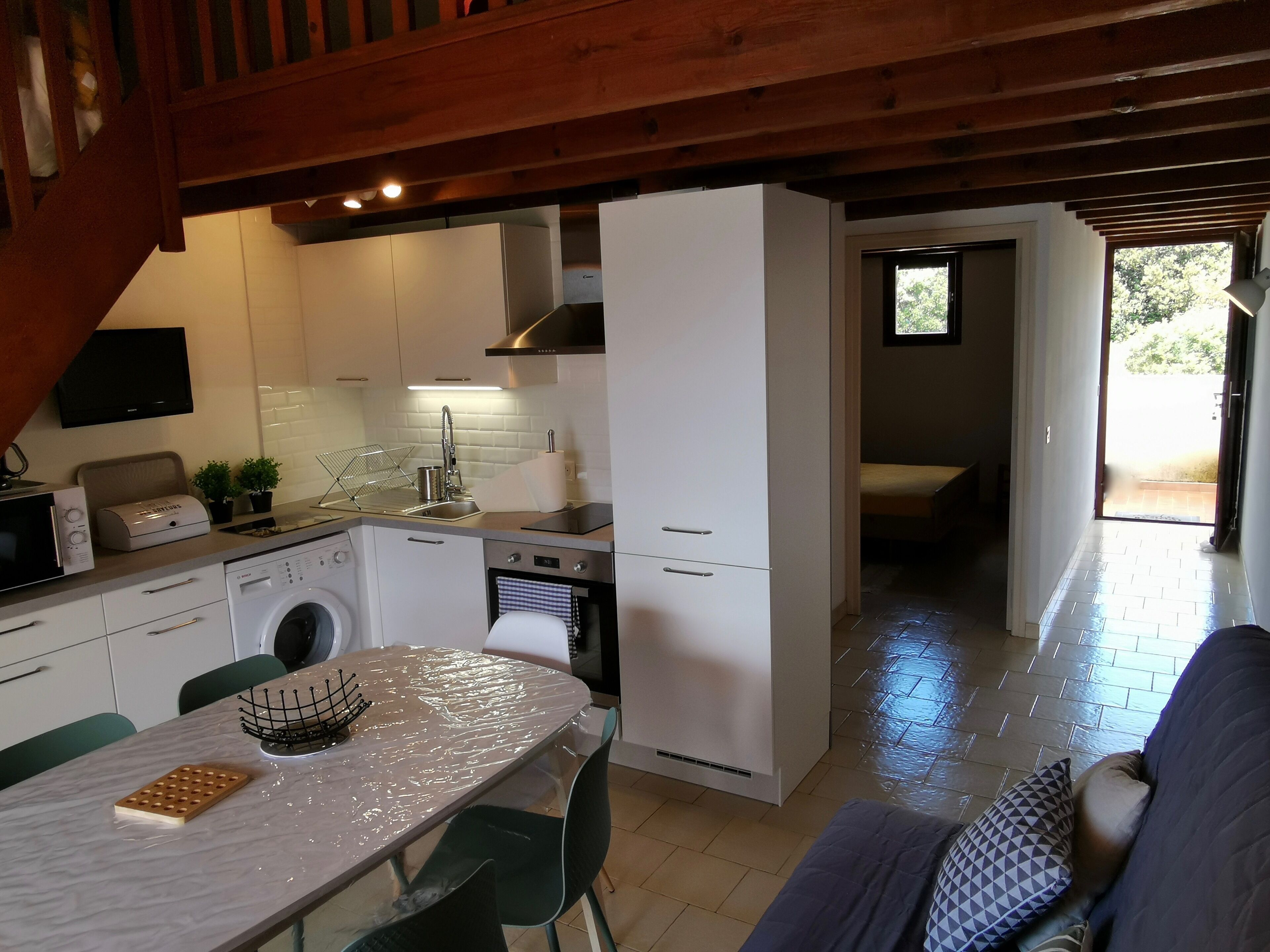 Een moderne keuken met een ingebouwde oven, magnetron en wasmachine. Er is een eethoek met een tafel en stoelen, en een bank in de woonkamer.