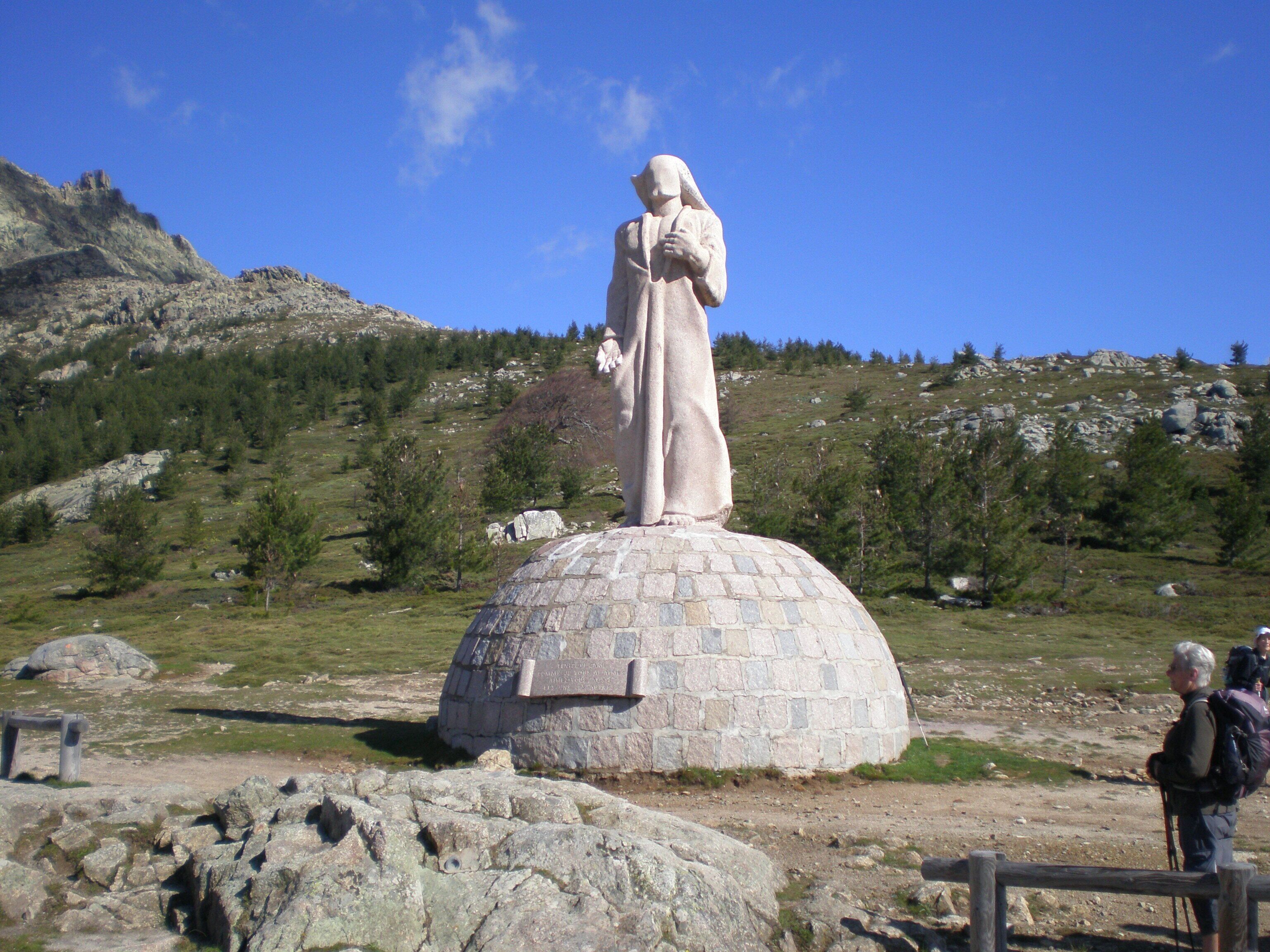 Une statue de pierre, posée sur un piédestal, dans un environnement naturel avec une chaîne de montagnes en arrière-plan.