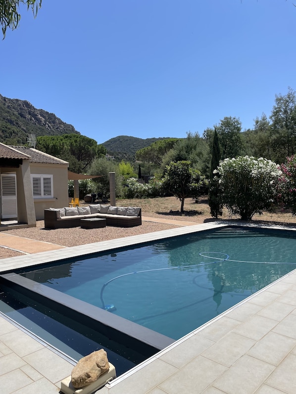 A swimming pool with a tiled deck, a sofa with cushions, and a small building with a tiled roof.