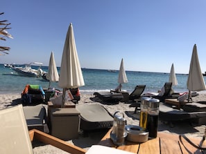 A beachside setting with lounge chairs, umbrellas, and a wooden table with a salt shaker and a bottle.