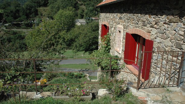 Ein Steinhäuschen mit roten Fensterläden, umgeben von viel Grün und einer hügeligen Landschaft.