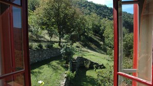 Blick aus einem Zimmer mit großem Fenster auf eine grüne Fläche mit Steinmauer und Baum.