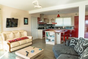 A modern living room with a beige sofa, a grey sofa, and a wooden coffee table. A kitchen with red cabinets and a dining area is visible in the background.