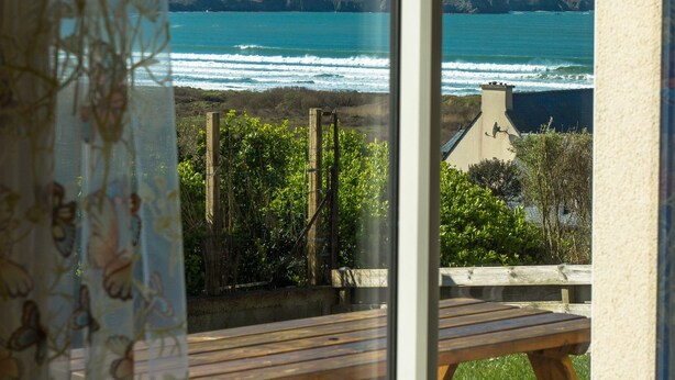 Vista dalla finestra su una spiaggia con onde, un pontile di legno e un edificio sullo sfondo.