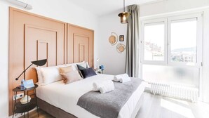 Et moderne soveværelse med en stor seng, et natbord, en lampe og udsigt over byen gennem vinduet.