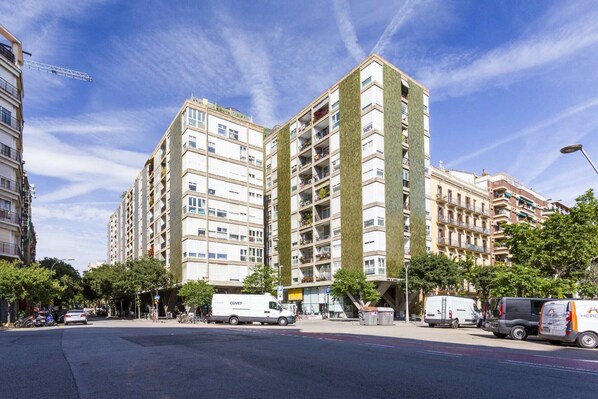 Un immeuble résidentiel à plusieurs étages, doté de balcons, d’un espace de stationnement et situé le long d’une rue où circulent des véhicules.