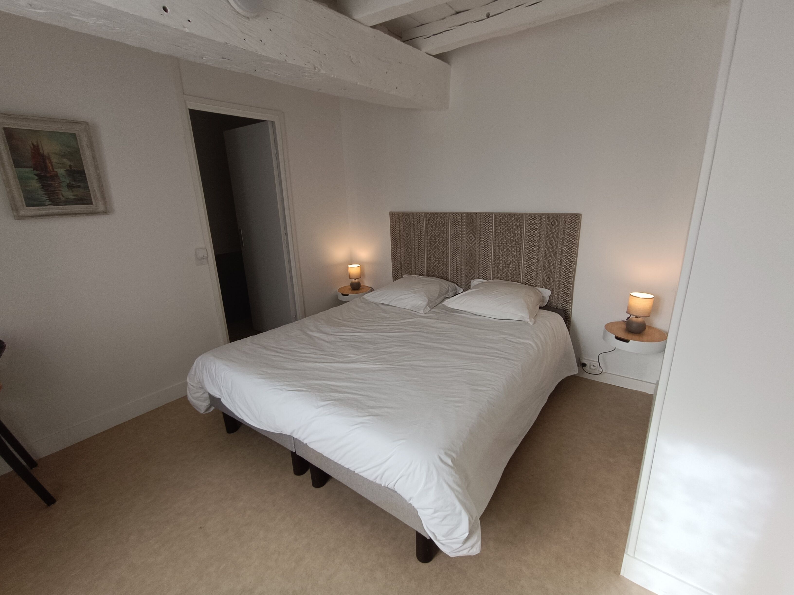 A bedroom with a bed, two bedside lamps, a door, and a painting on the wall.