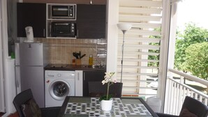 A compact kitchen with a washing machine, a microwave, and a coffee maker. There is a dining area with a table and chairs, and a balcony with a view of trees. - Promotions 280th week Beautiful apartment StFrançois Domaine Anse des Rocher (97180)