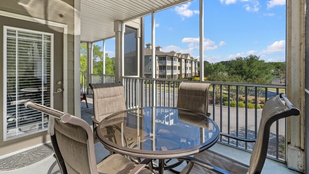 A covered outdoor area with a glass table and chairs, a ceiling fan, and a view of a multi-story building. - 1115 Crow Creek Drive (Calabash)