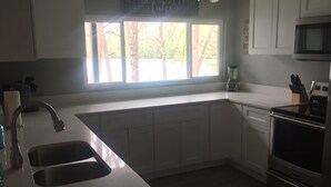 Eine Küche mit weißen Schränken, einer Edelstahl-Spüle, einem Fenster mit Blick auf Bäume und einer Mikrowelle.