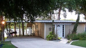 Ein kleines, eingeschossiges Gebäude mit überdachter Terrasse, Eingangstür und einem kleinen Garten mit Baum und Sträuchern.
