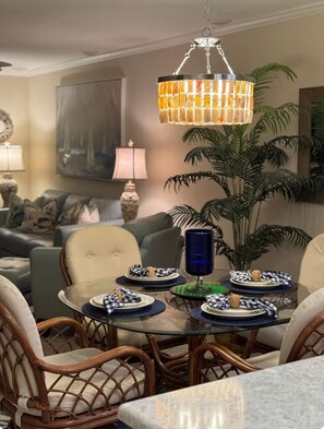 A dining area with a round table, wicker chairs, a hanging chandelier, and a potted plant. - Elegant Vero on the Island (Vero Beach)