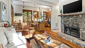 A spacious living area with a stone fireplace, a television mounted above it, and a kitchen in the background. - Owl's Nest - 2/2.5, Luxurious, Pet Friendly Home (Black Mountain)