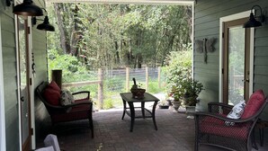 A covered porch with a ceiling fan, two wicker chairs with cushions, a small table with a potted plant, and a brick floor.
