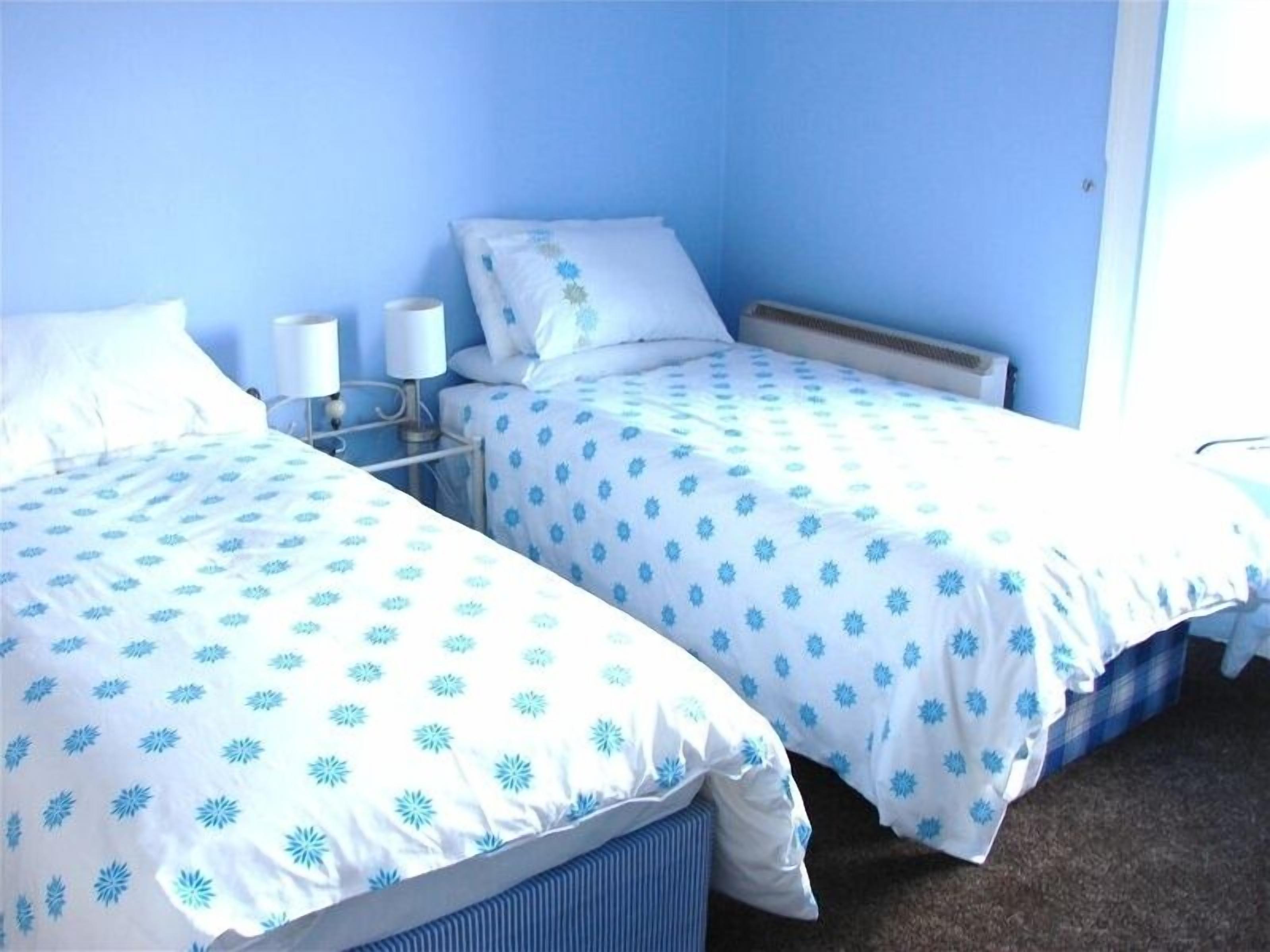A room with two single beds, each with a blue floral bedspread and white pillows. There are bedside tables with lamps and a small shelf.