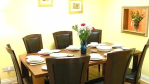 A dining room with a wooden table set for six, surrounded by six chairs. There are framed pictures on the wall above the table.