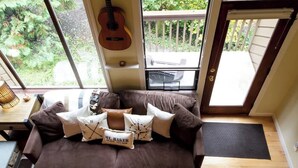 Ein braunes Sofa mit vielen Kissen, eine Gitarre an der Wand und Blick ins Grüne.