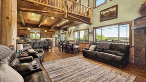 A spacious living area with a high ceiling, wooden beams, a fireplace, and large windows offering views of the outdoors.