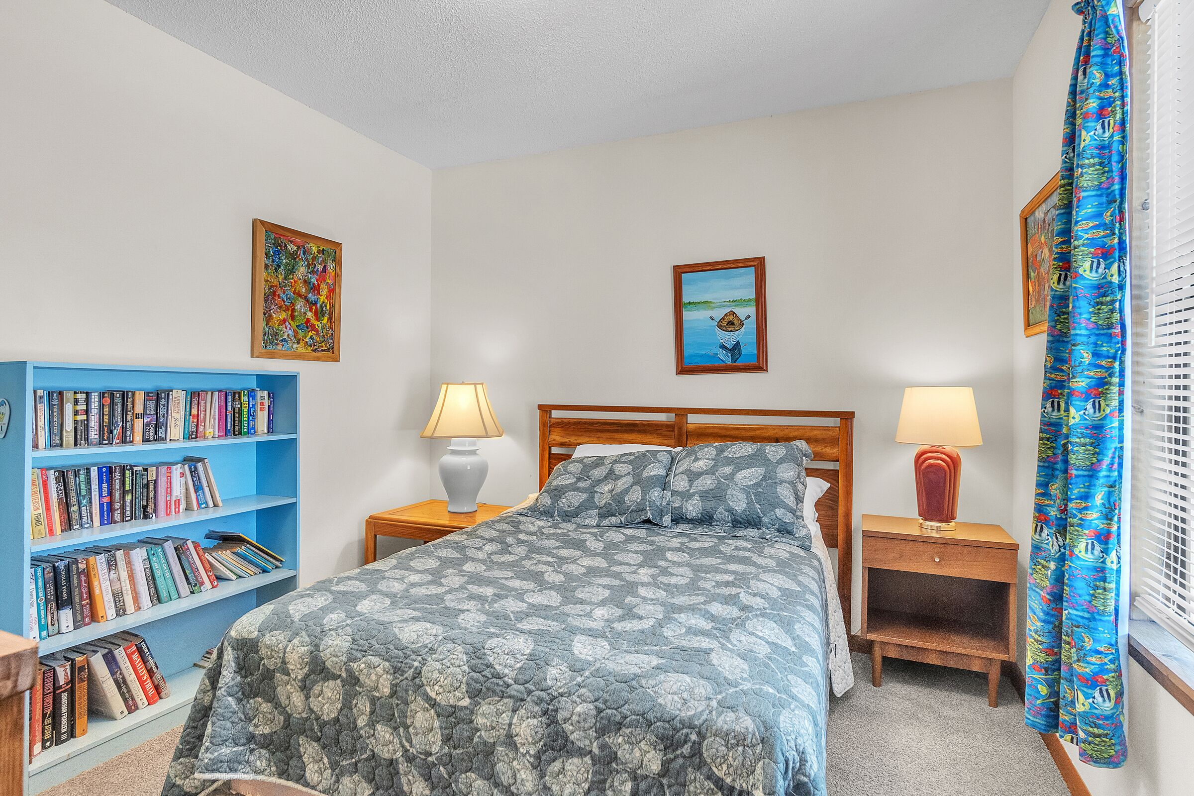 A bedroom with a bed, a bookshelf, two bedside tables with lamps, and a window with curtains.