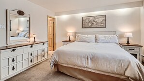 A bedroom with a large bed, bedside tables with lamps, a dresser, a world map on the wall, and a door leading to another room.