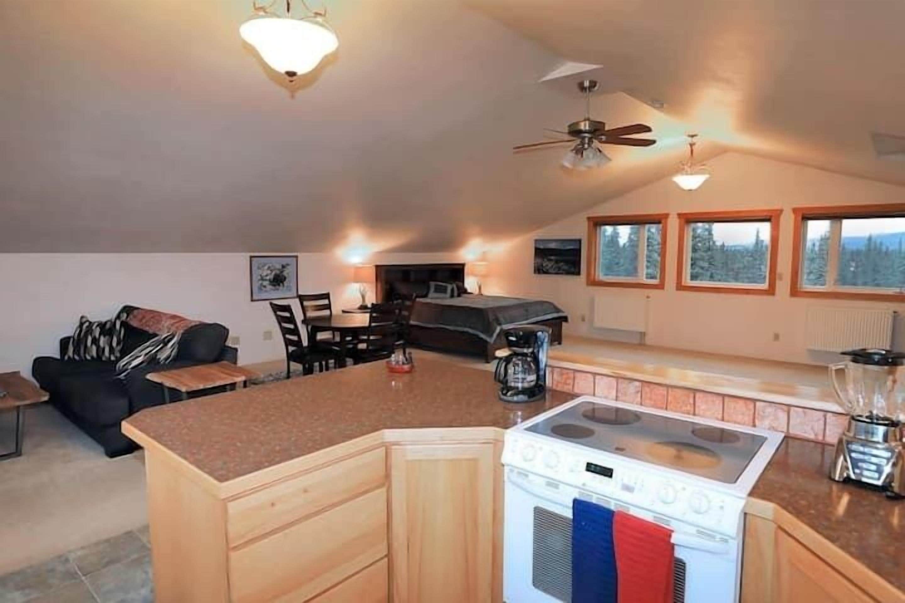 Une cuisine dotée d’un îlot central, d’une cuisinière et d’un mixeur. Des fenêtres offrent une vue sur les arbres et un ventilateur de plafond est installé au plafond.