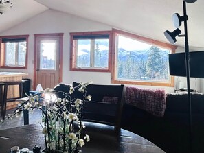 Un salon chaleureux avec une table en bois, un canapé noir et une grande fenêtre donnant sur les montagnes.