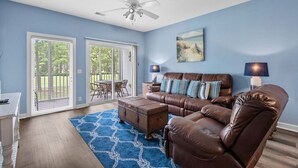 A living room with a brown leather sofa, a coffee table, and a blue patterned rug. There is a ceiling fan, a painting on the wall, and a patio with a table and chairs visible through the glass doors. - 3709 Champions at Sea Trail (Sunset Beach)