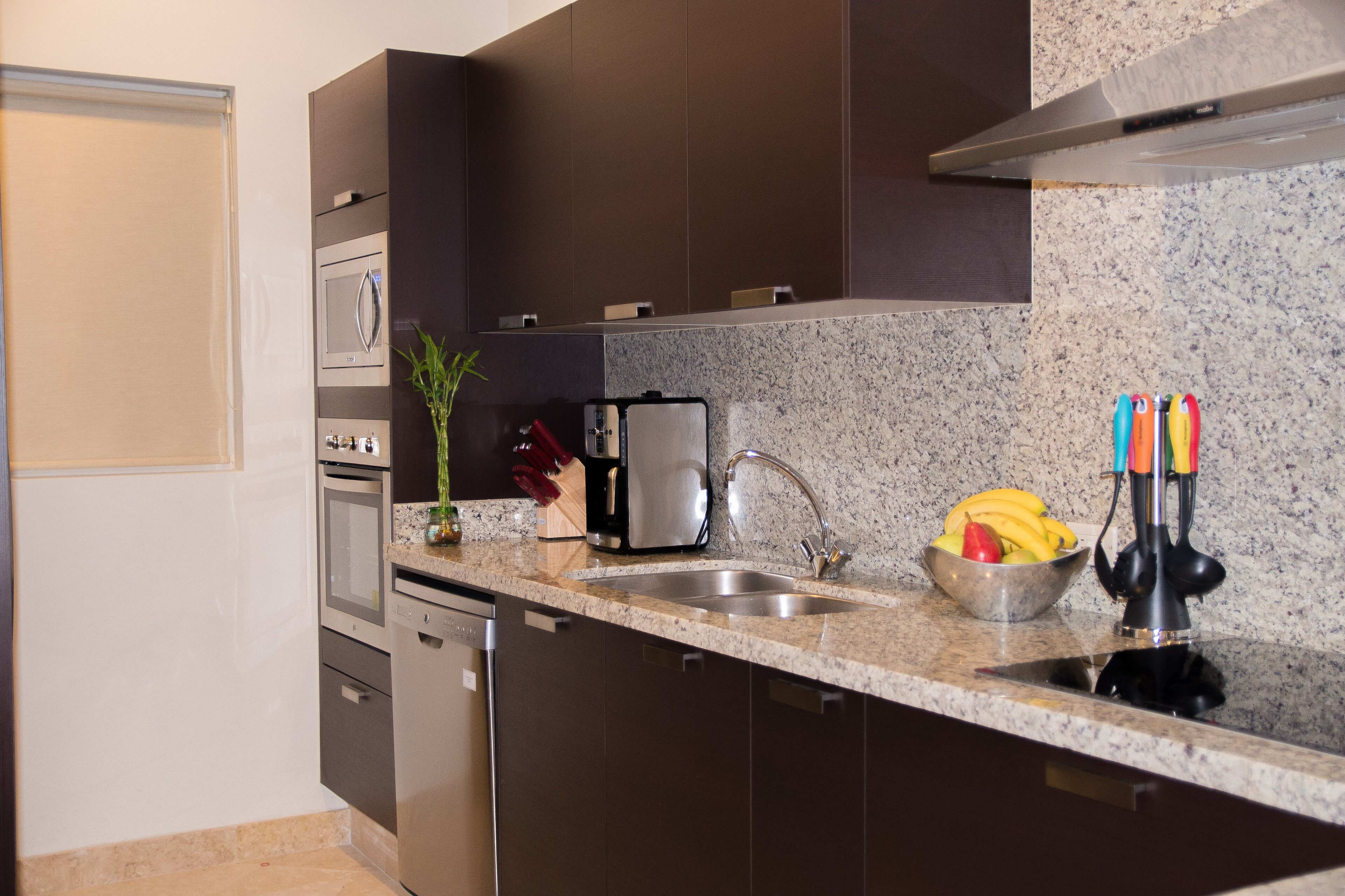 Een moderne keuken met donkere kastjes, een granieten aanrechtblad, een roestvrijstalen spoelbak, een magnetron, een koffiezetapparaat en een schaal fruit.