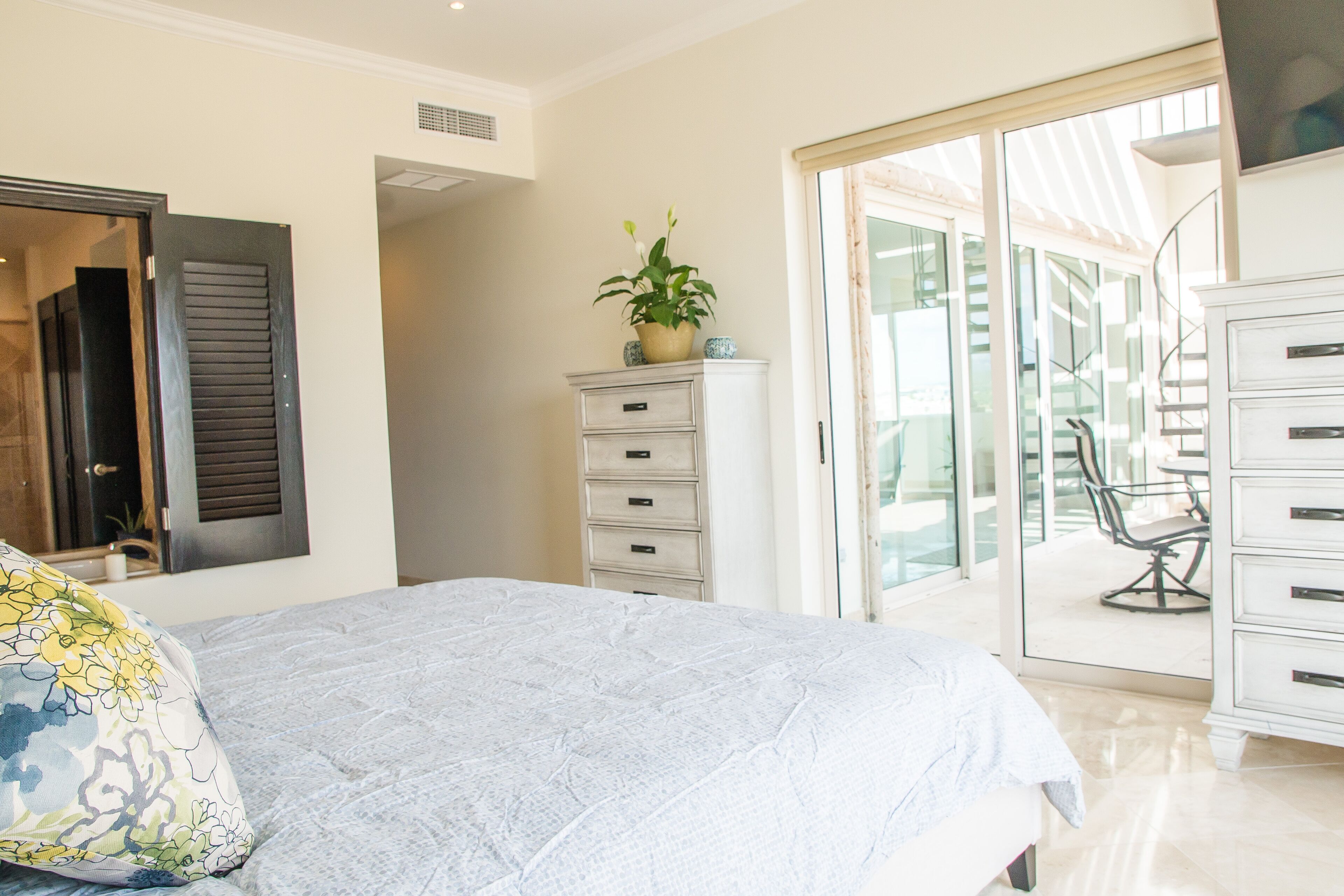 Une chambre à coucher comprenant un lit, une commode, une plante et une porte-fenêtre coulissante donnant sur un balcon.