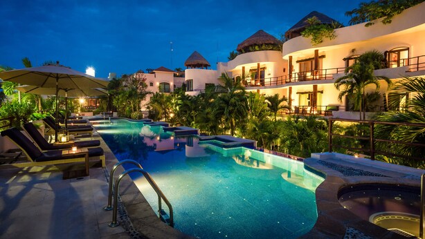 A swimming pool with lounge chairs and umbrellas, surrounded by palm trees and buildings with thatched roofs. - Steps from Beach,  Large private balcony overlooking pool & tropical garden (Playa del Carmen)
