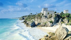A coastal scene with a beach, clear blue water, and ancient stone structures on a cliff. - Steps from Beach,  Large private balcony overlooking pool & tropical garden (Playa del Carmen)