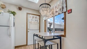 A compact kitchen with a white refrigerator, a wooden table with matching chairs, a hanging chandelier, and a window with a view of the outside.