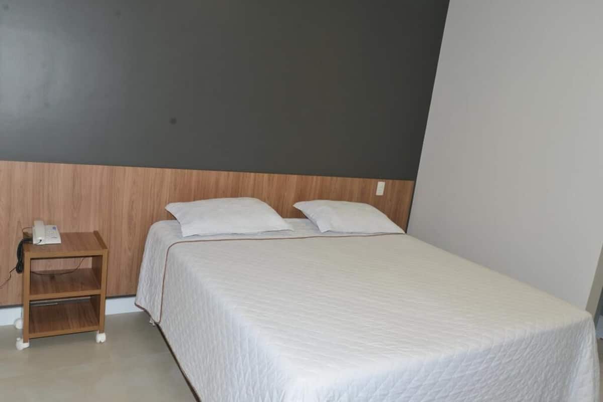 A single bed with a white quilted bedspread and two pillows, a wooden nightstand with a phone, and a plain wall with a dark panel above the bed.