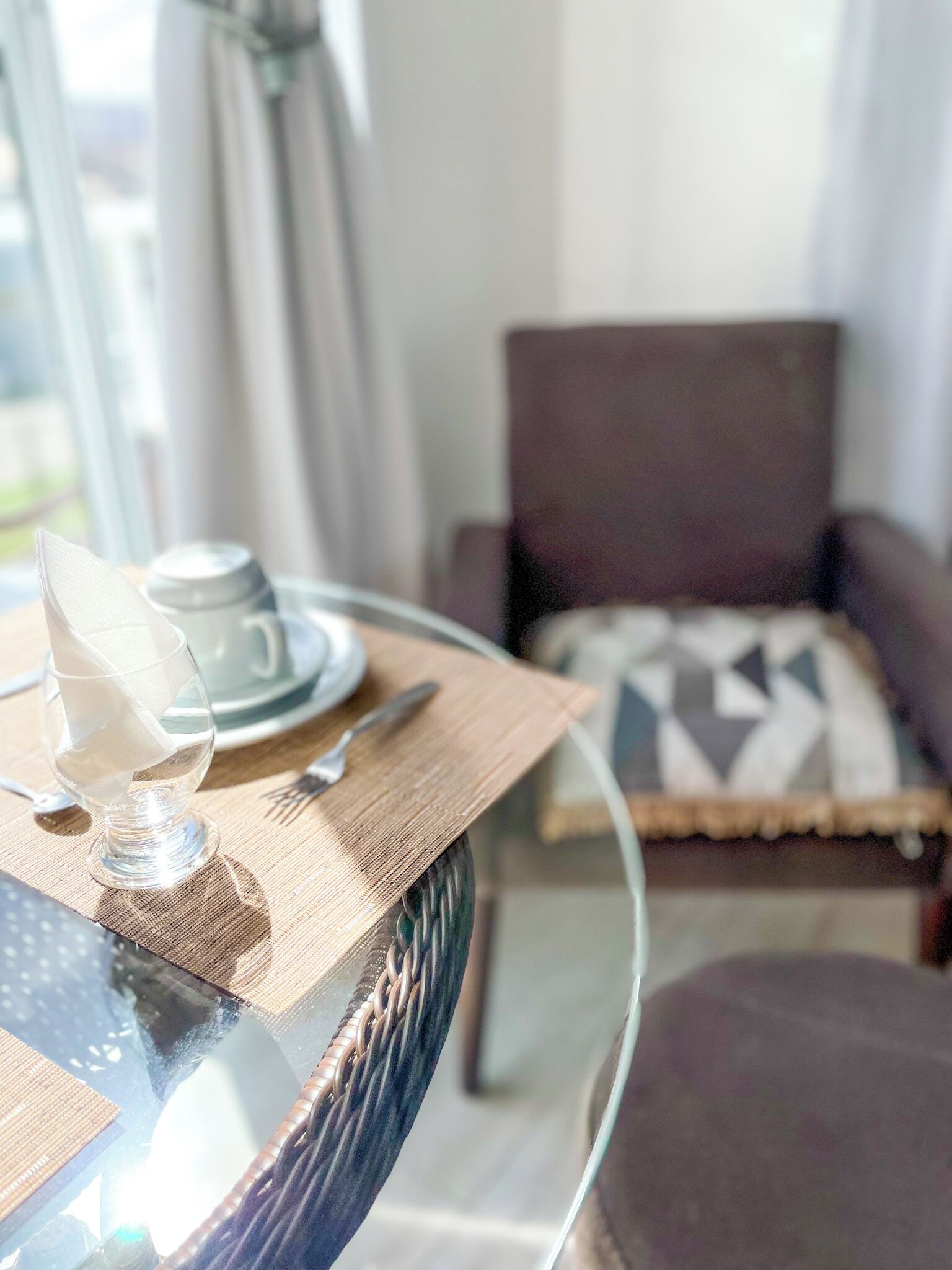 A dining table with a glass top, set with a white napkin, a glass, a plate, and cutlery. A chair with a geometric cushion is in the background.