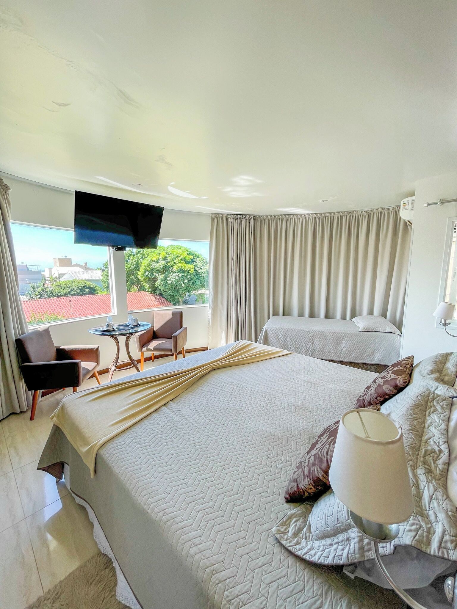 A hotel room with a large bed, a smaller bed, a TV, a table with chairs, and a view of the outdoors.