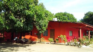 A red building with a covered porch, surrounded by greenery and a few potted plants. - Hospedaje Yaquelin (Cambutal)