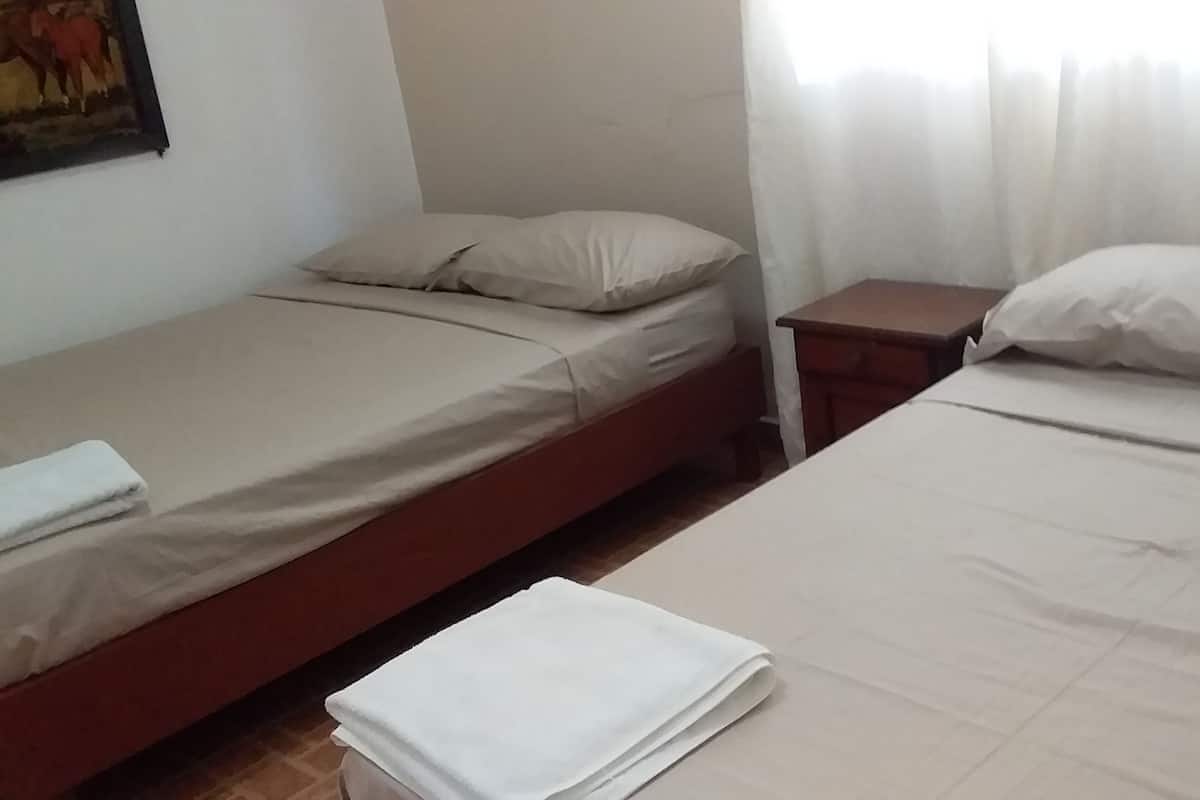 Ein Zimmer mit zwei Einzelbetten, jeweils mit Kissen und Decke. Dazwischen steht ein kleiner Nachttisch.