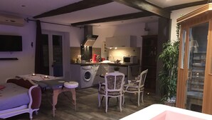 A cozy living area with a sofa, a small table, and a chair. There is a washing machine and a microwave in the kitchen area. A bathtub is visible in the foreground. - Le Mas Romantique (Senas)