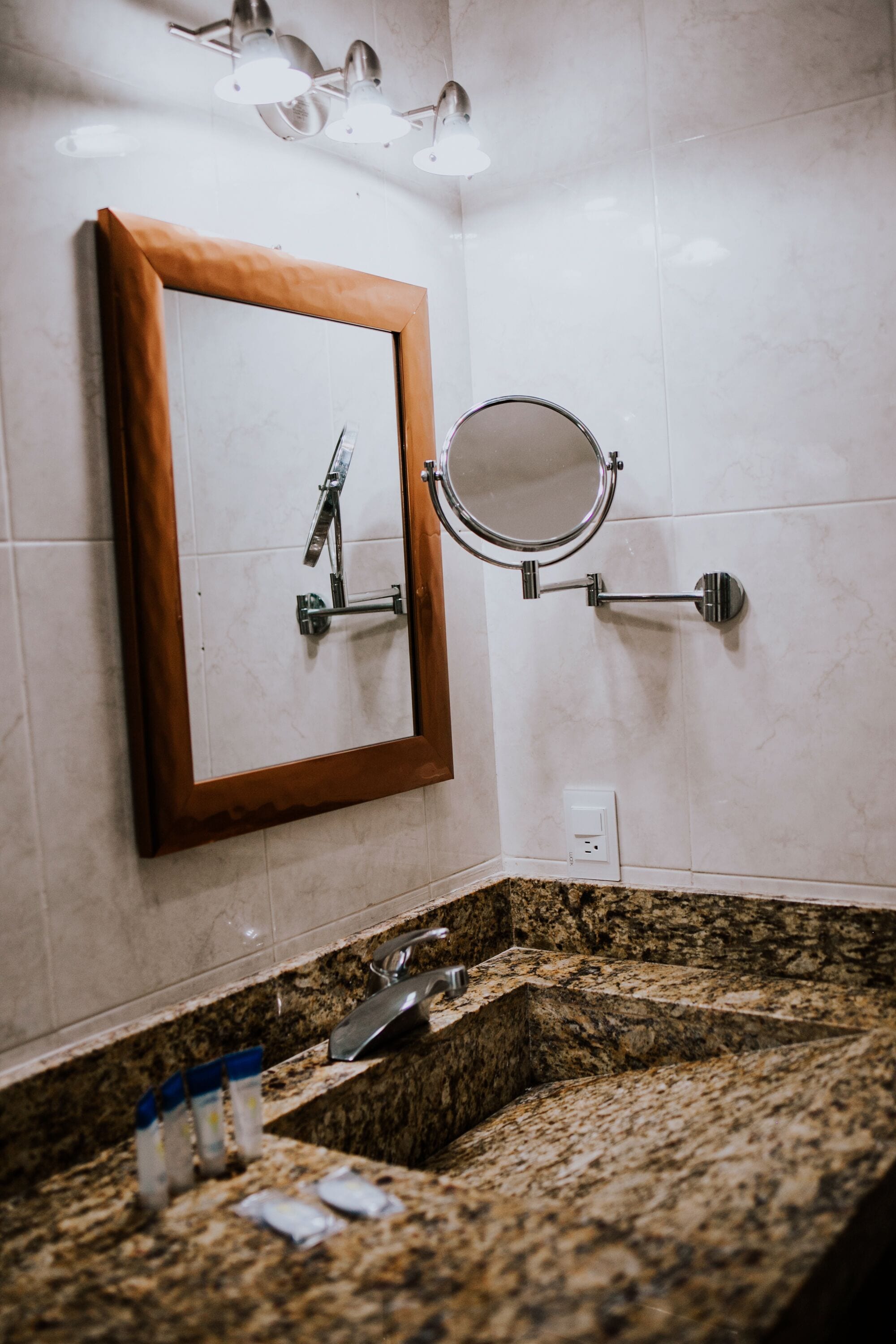 A bathroom with a granite countertop, a wooden-framed mirror, a round mirror, and a showerhead.