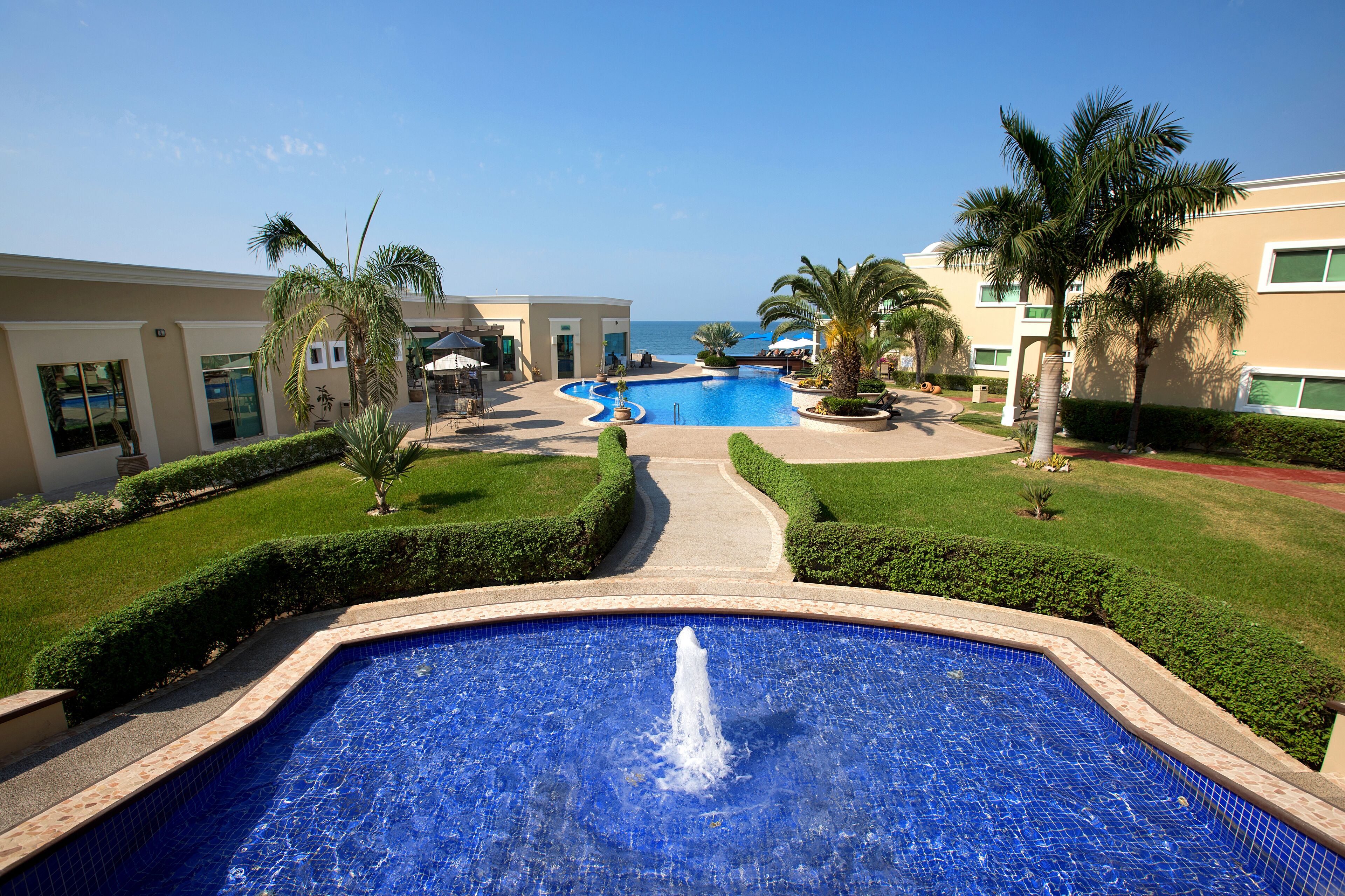 A swimming pool with a fountain, surrounded by well-maintained lawns and palm trees, with a view of the ocean in the background.