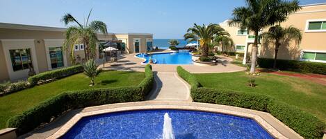 A swimming pool with a fountain, surrounded by well-maintained lawns and palm trees, with a view of the ocean in the background.