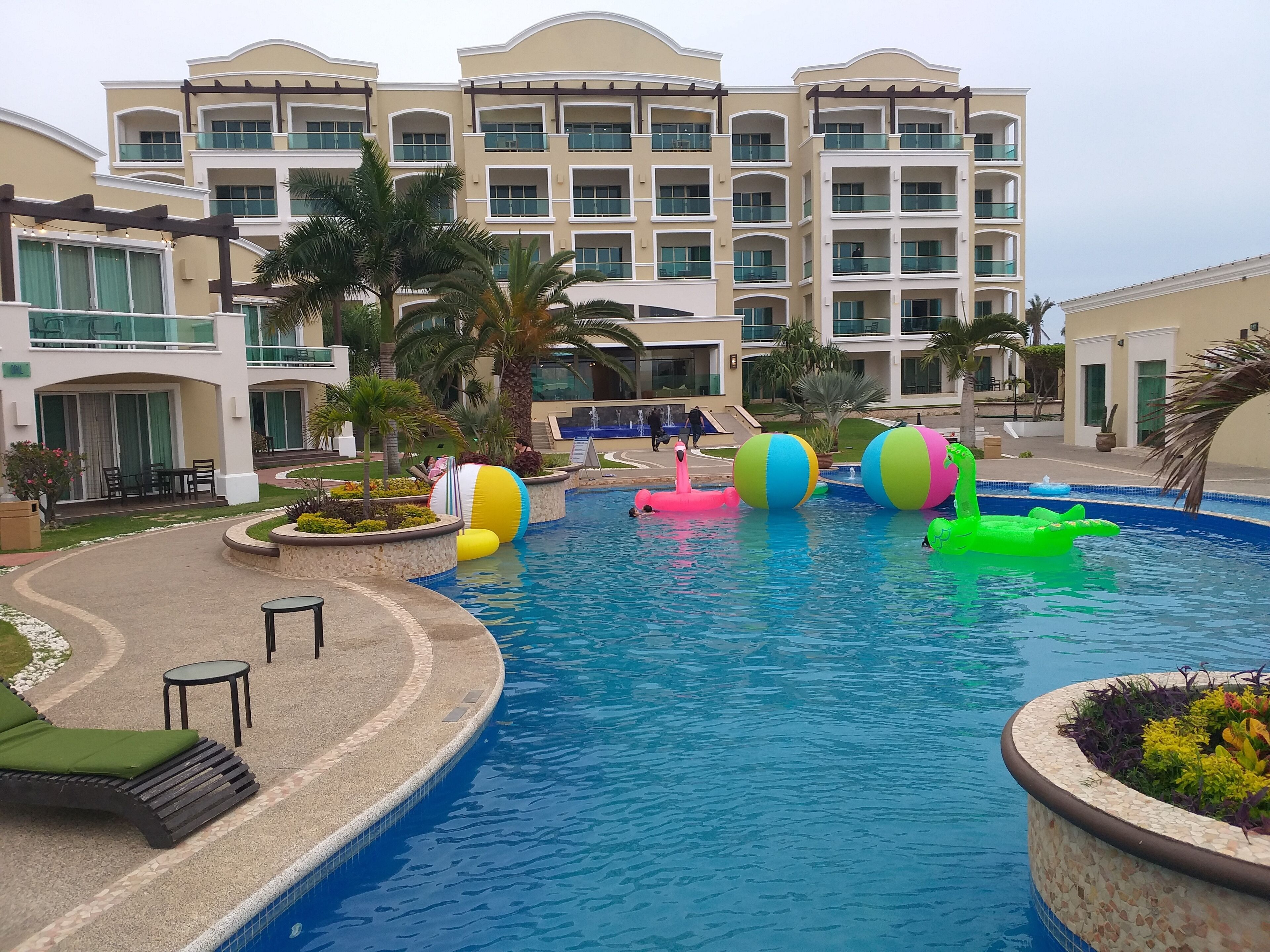 Une piscine avec des jouets gonflables de couleurs vives, un espace détente abrité et un immeuble à plusieurs étages doté de balcons.