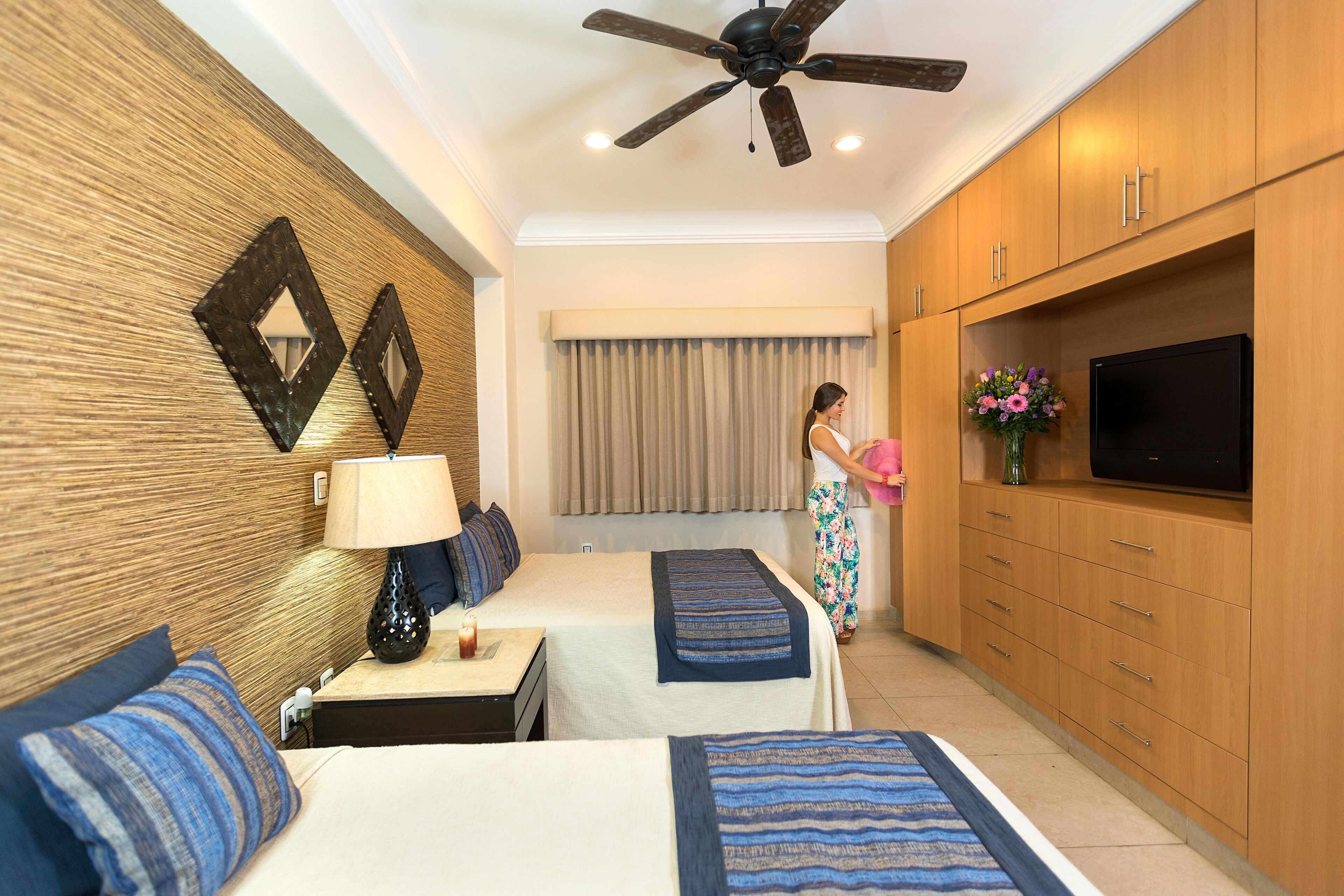 A hotel room with two beds, a ceiling fan, wooden cabinets, a TV, and a window with curtains.